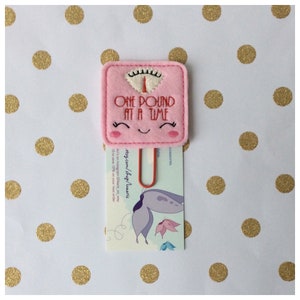 May include: A pink felt scale-shaped paper clip with the text "ONE POUND AT A TIME" embroidered in red. The clip has a smiling face design and holds a card with a butterfly illustration and shop details.