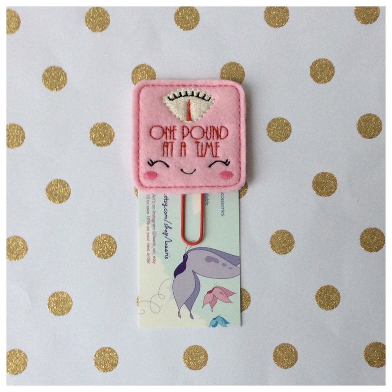 May include: A pink felt scale-shaped paper clip with the text "ONE POUND AT A TIME" embroidered in red. The clip has a smiling face design and holds a card with a butterfly illustration and shop details.
