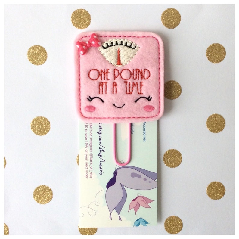 May include: A pink felt bookmark shaped like a scale with the text "ONE POUND AT A TIME". The bookmark has a smiling face and a small bow. A pink paper clip holds a card with a butterfly illustration.
