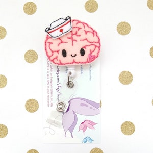 Op de afbeelding: Een roze viltbrein met een witte verpleegstersmuts en een rood kruis. De hersenen hebben een glimlachend gezicht en zijn bevestigd aan een transparante, intrekbare badgehouder van plastic. De badgehouder is bevestigd aan een witte kaart met de tekst "Accessories available", "Clips", "u.etsy.com/shop/LaAri", "LaAri's on Instagram @laaris_on_etsy", en "SAVE 10 to save 10% on your next order".