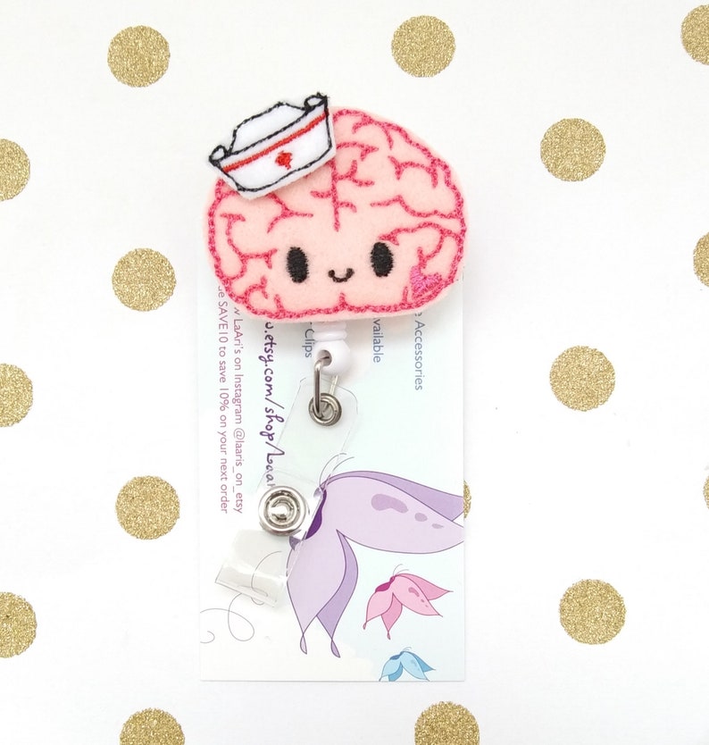 Op de afbeelding: Een roze viltbrein met een witte verpleegstersmuts en een rood kruis. De hersenen hebben een glimlachend gezicht en zijn bevestigd aan een transparante, intrekbare badgehouder van plastic. De badgehouder is bevestigd aan een witte kaart met de tekst "Accessories available", "Clips", "u.etsy.com/shop/LaAri", "LaAri's on Instagram @laaris_on_etsy", en "SAVE 10 to save 10% on your next order".