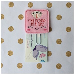 May include: A pink felt bookmark shaped like a scale, with the text "ONE POUND AT A TIME" embroidered in red. The scale has a smiling face and is attached to a metal clip. The background is white with gold polka dots.