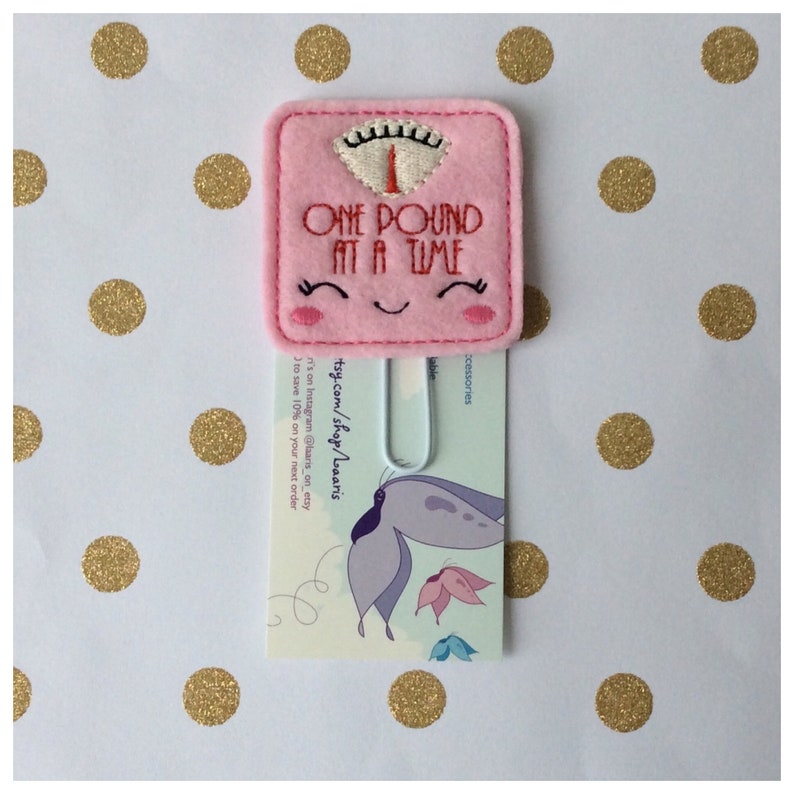 May include: A pink felt bookmark shaped like a scale, with the text "ONE POUND AT A TIME" embroidered in red. The scale has a smiling face and is attached to a metal clip. The background is white with gold polka dots.