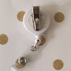 Op de afbeelding: Witte intrekbare badgehouder met een zilveren clip en een transparante plastic band. De houder bevindt zich op een witte achtergrond met gouden stippen.