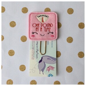 May include: A pink felt scale-shaped paper clip with the text "ONE POUND AT A TIME" embroidered in red. The scale has a smiling face and is attached to a card with a gold paper clip. The background is white with gold polka dots.