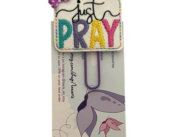 May include: A white square embroidered patch with the black cursive text just and the colorful block letters PRAY, featuring a small purple polka dot bow at the top left corner, attached to a purple metal paperclip on a branded display card.