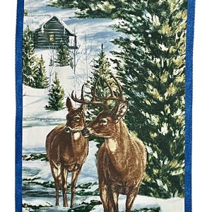 May include: A fabric wall hanging depicting a winter scene with two deer in a snowy landscape. A cabin is among evergreen trees. The border is a solid blue.