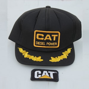Könnte beinhalten: Schwarze Trucker-Kappe mit einem gelben "CAT DIESEL POWER" Logo und goldenen, gestickten Blättern am Schirm. Ein separates gelbes "CAT" Logo-Patch ist ebenfalls enthalten.
