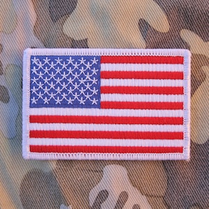 May include: Embroidered rectangular patch featuring the American flag. The flag has a blue field with white stars and red and white stripes. The patch is bordered with white stitching and set against a camouflage background.