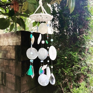 Puede incluir: Un móvil eólico hecho a mano con una parte superior de macramé y discos de concha iridiscentes colgantes, cuentas y detalles de cristal. El móvil eólico está suspendido de una rama de árbol, con una borla verde y un prisma de cristal transparente. La combinación de colores es blanca, transparente y verde.