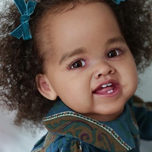 May include: A close-up of a smiling baby doll with brown eyes and dark curly hair. The doll is wearing a blue and brown patterned dress with a white collar and has two teal velvet bows in her hair.