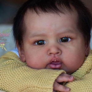 May include: Close-up portrait of a baby doll with brown eyes and a light brown complexion. The doll is wearing a yellow knitted sweater.