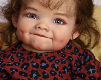 May include: A close-up of a baby doll with brown curly hair, blue eyes, and rosy cheeks. The doll is wearing a red and blue leopard print dress with a blue bow in her hair.