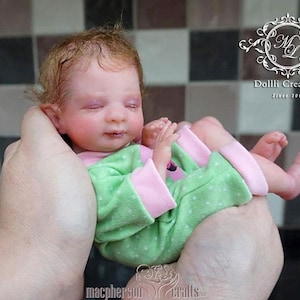 Peut inclure: Une petite poupée reborn endormie avec les yeux fermés et une tenue à pois rose et vert. La poupée est tenue dans les mains de quelqu'un.