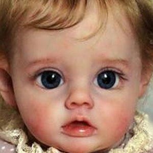May include: A close-up of a baby doll with blue eyes and rosy cheeks. The doll is wearing a pink and white polka dot dress with a lace trim.