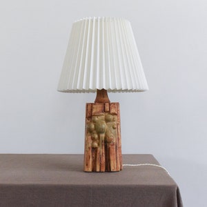 May include: A ceramic table lamp with a white pleated lampshade. The base of the lamp is textured and features a geometric design with brown, green, and yellow accents.