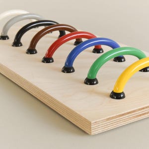 May include: A wooden board with seven colourful plastic handles attached. The handles are in the shape of an arch and are arranged in a row from left to right. The colours of the handles are white, grey, black, brown, red, blue, and yellow.