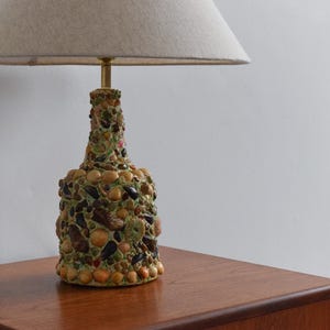 May include: A table lamp with a beige lampshade and a base made of a bottle covered in mosaic tiles. The tiles are various shades of brown, green, and yellow.