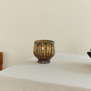 May include: A ceramic cup with a brown base and a tan body with dark brown drips. The cup sits on a white tablecloth. A wooden chair and a wooden bowl are also visible.