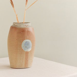 May include: A tan ceramic vase with a light blue circular design and vertical lines. The vase has a wide base and a narrower neck, with dried grasses inside. The vase is on a white surface.