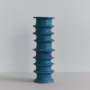 May include: A tall, teal-colored ceramic vase with a unique, ridged design. The vase has a cylindrical shape with stacked, rounded ridges. The base is a reddish-brown color. The vase is approximately 12 inches tall.