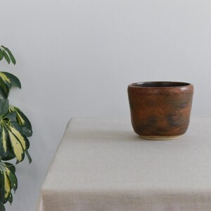 Peut inclure: Un petit pot en céramique marron fait à la main avec une surface texturée. Le pot cylindrique a un bord légèrement évasé et repose sur une base de couleur claire. Il est présenté sur une nappe de couleur neutre.