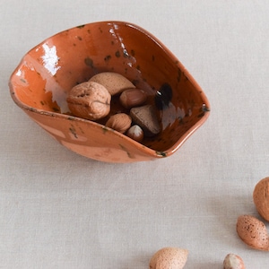 May include: A rustic, terracotta-coloured bowl filled with a variety of nuts, including almonds, walnuts, and hazelnuts. The bowl has a unique, uneven shape and a speckled glaze.