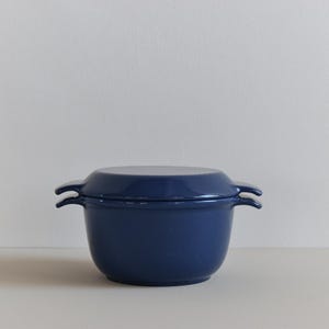 May include: A navy blue cast iron Dutch oven with a lid. The pot has two loop handles. It is on a white surface, with a plain white wall in the background. The pot is suitable for cooking.