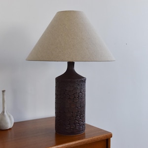 May include: A dark brown ceramic table lamp with a textured, cylindrical base and a beige lampshade. The lamp is on a wooden table, with decorative vases in the background. The lamp is approximately 61 cm tall.