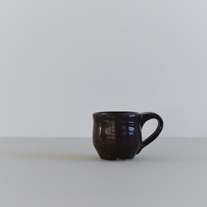 May include: A small, dark brown ceramic mug with a rounded body and a curved handle. The mug has a textured surface and appears to be handmade. The mug is sitting on a light-coloured surface.