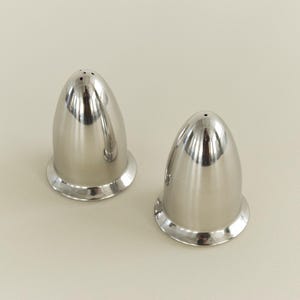 May include: Two shiny, silver-colored salt and pepper shakers. Each shaker has a rounded, egg-like shape with a flared base. The top of each shaker has small holes for dispensing the contents. The shakers are on a light beige surface.