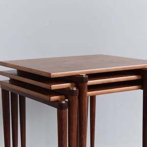 May include: Three wooden nesting tables with square tops and dark brown legs. The tables are stacked, revealing their different sizes. The wood has a warm, natural finish, and the design is mid-century modern. The tables are approximately 50 cm in height.