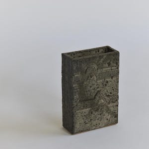 May include: A dark grey ceramic vase with a textured surface and a geometric pattern. The vase is rectangular in shape and has a wide opening.