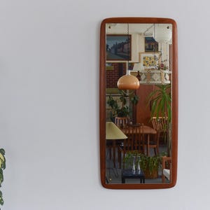 May include: A rectangular mirror with a wooden frame, reflecting a room with vintage furniture and decor. The mirror's frame has rounded corners and a warm, brown tone. The reflection shows a dining table, artwork, and plants, creating a retro aesthetic.