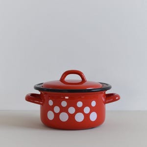 May include: A bright red enamel pot with a matching lid, featuring a pattern of white polka dots of varying sizes. The pot has two side handles and a black rim around the top edge and the lid, providing a clear contrast to the red surface.