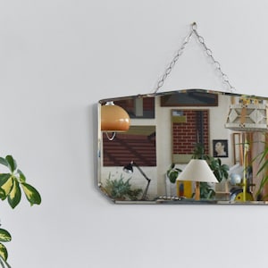May include: A rectangular, vintage-style mirror with a unique, geometric shape, hanging from a silver chain. The mirror reflects a room with various objects, including lamps, artwork, and plants. The mirror's edges are beveled, adding to its retro aesthetic.