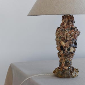 Peut inclure: Une lampe de table avec un abat-jour en tissu beige et une base en petites pierres de couleur brun, beige et noir.