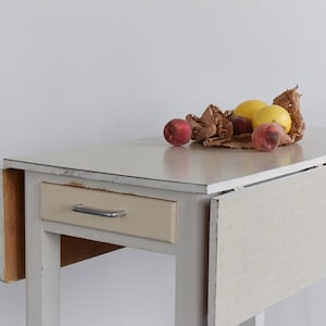 May include: A white, vintage drop-leaf table with a beige drawer and a silver handle. The table surface holds a collection of fresh fruit, including peaches and lemons. The table is set against a plain white background.