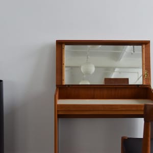 May include: A teak wood vanity desk featuring a flip-top lid that reveals a rectangular mirror. The desk has a light-coloured surface and is accompanied by a wooden chair with a black seat. A black cylindrical stand holds a vase and books.