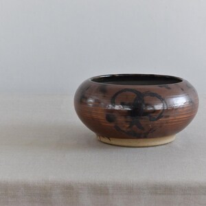 May include: A handcrafted, round ceramic bowl with a dark brown exterior and a lighter beige base. The bowl features a black, abstract design on the front. The interior is dark. A decorative piece for the home.