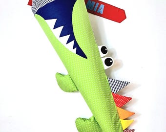 May include: A green, cone-shaped school bag designed as an alligator. The bag features a gingham top, a red ribbon, and a name tag that says "MIA". The alligator design includes white teeth, googly eyes, and colorful triangular accents.