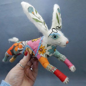May include: A handmade white rabbit plush toy with long ears, wearing small round glasses. The rabbit's body is adorned with colorful floral fabric, and it has a small, fluffy tail. The toy is held against a blue background.