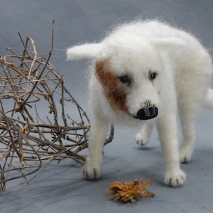 Puede incluir: Escultura artesanal de un perro blanco con una mancha marrón en la cara y una nariz negra. El perro está cerca de un montón de ramitas y una pequeña flor seca. La escultura está hecha de lana.