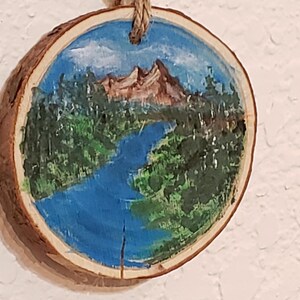 May include: A circular wooden slice featuring a hand-painted landscape. The artwork shows a mountain, river, and trees under a blue sky. A natural rope is attached for hanging. The wood slice has a natural bark edge.