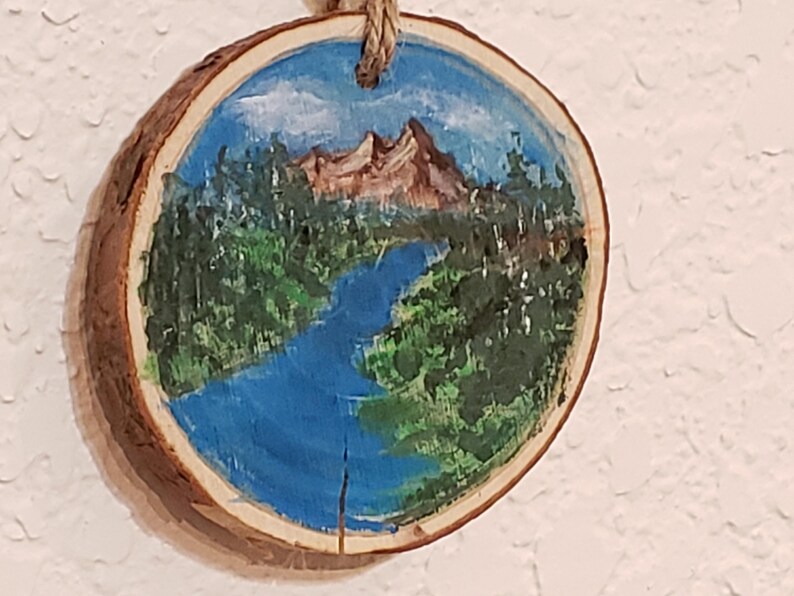 May include: A circular wooden slice featuring a hand-painted landscape. The artwork shows a mountain, river, and trees under a blue sky. A natural rope is attached for hanging. The wood slice has a natural bark edge.