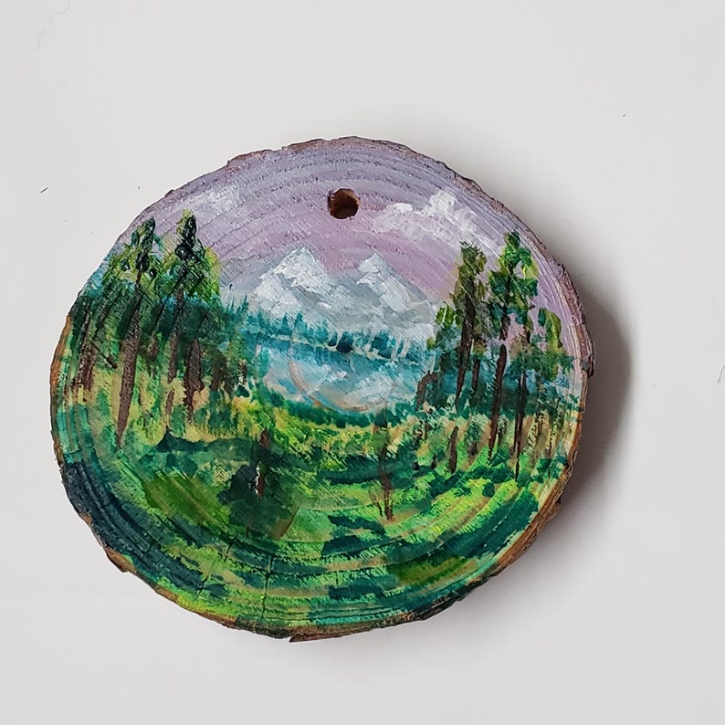 May include: A hand-painted landscape on a wooden slice. The artwork shows a mountain range, trees, and a lake, using green, blue, purple, and white. The wood slice has a natural bark edge and a hole at the top for hanging.