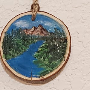 May include: A circular wooden decoration featuring a painted landscape. The image shows a blue river flowing through green trees, with brown mountains and a blue sky. A natural twine loop is attached for hanging.