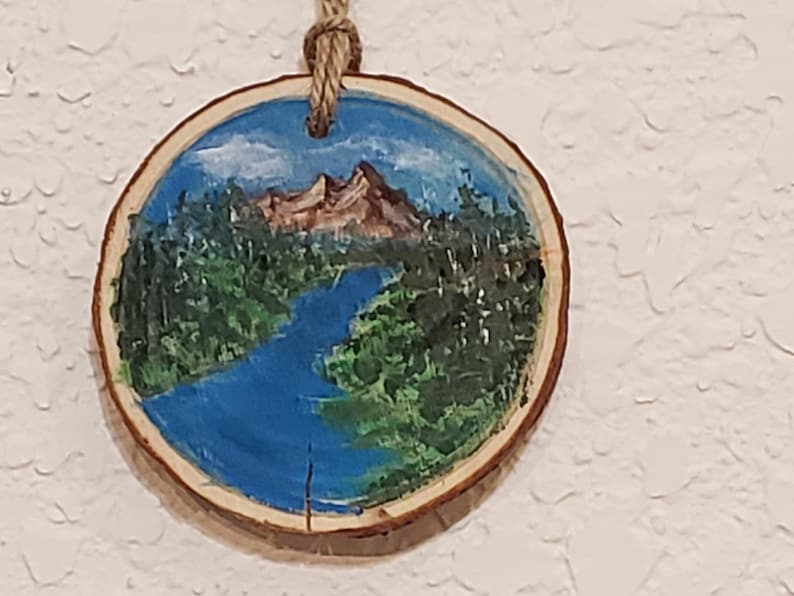 May include: A circular wooden decoration featuring a painted landscape. The image shows a blue river flowing through green trees, with brown mountains and a blue sky. A natural twine loop is attached for hanging.