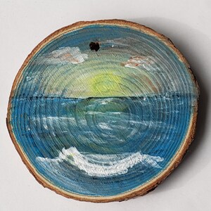 May include: A circular wooden slice with a painted ocean scene. The artwork shows a sunset over the sea, with blue and white waves, a yellow and green sun, and clouds. The wood has a natural, brown edge and a hole at the top for hanging.