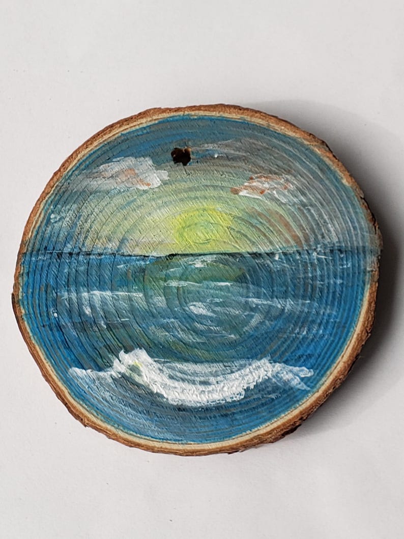 May include: A circular wooden slice with a painted ocean scene. The artwork shows a sunset over the sea, with blue and white waves, a yellow and green sun, and clouds. The wood has a natural, brown edge and a hole at the top for hanging.